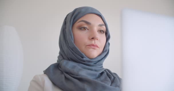 Closeup portrait of young muslim attractive female doctor in hijab and white coat typing on the laptop looking at camera smiling cheerfully sitting in the armchair in the white room indoors - Video, Çekim