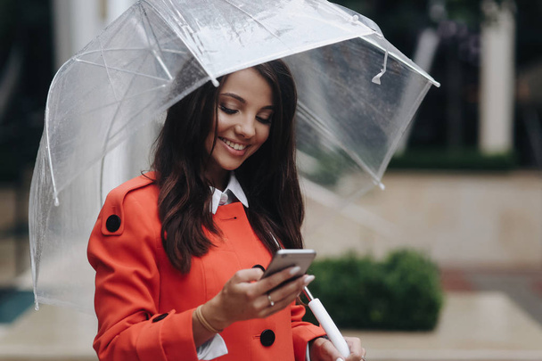 Beautiful smiling woman with umbrella rainy day in red coat looking attentively at screen of cellphone and standing near office center. - Φωτογραφία, εικόνα