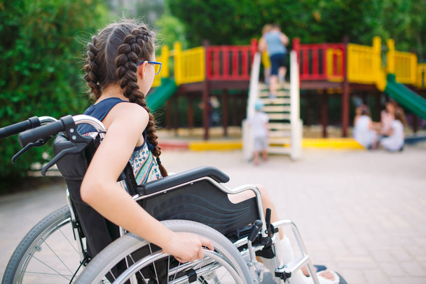 A girl with a broken leg sits in a wheelchair in front of the playground - Foto, afbeelding