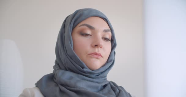 Closeup portrait of young muslim attractive female doctor in hijab and white coat typing on the laptop looking at camera sitting in the armchair in the white room indoors - Imágenes, Vídeo