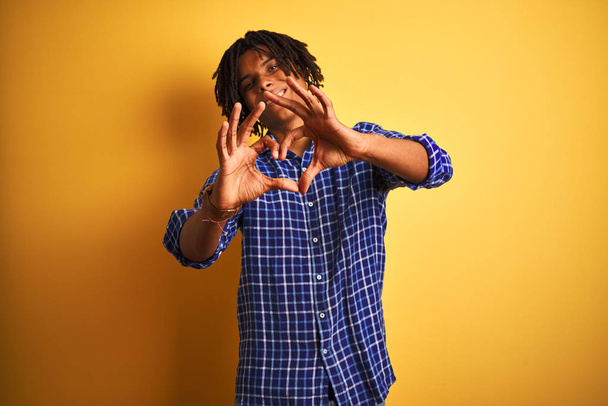 Afro man with dreadlocks wearing casual shirt standing over isolated yellow background smiling in love doing heart symbol shape with hands. Romantic concept. - Foto, afbeelding