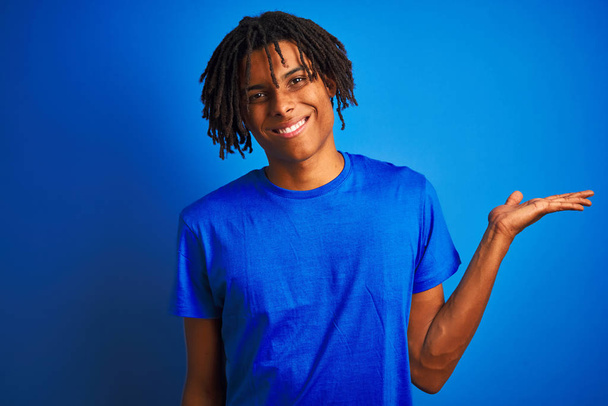 Afro american man with dreadlocks wearing t-shirt standing over isolated blue background smiling cheerful presenting and pointing with palm of hand looking at the camera. - Zdjęcie, obraz