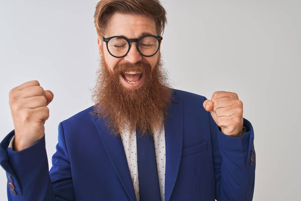 Young redhead irish businessman wearing suit and glasses over isolated white background very happy and excited doing winner gesture with arms raised, smiling and screaming for success. Celebration concept. - Photo, Image