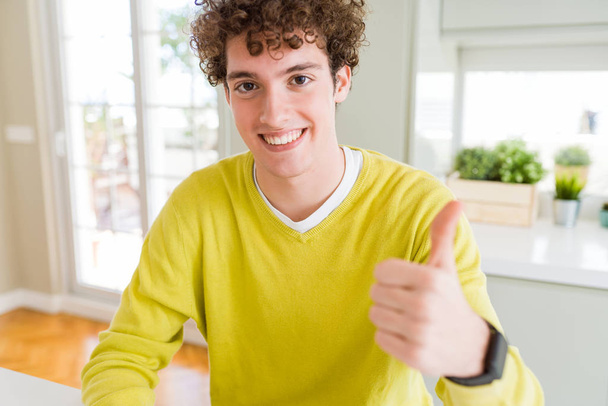 Joven hombre guapo usando suéter amarillo en casa haciendo un gesto feliz pulgares hacia arriba con la mano. Aprobación de la expresión mirando a la cámara mostrando éxito
. - Foto, imagen
