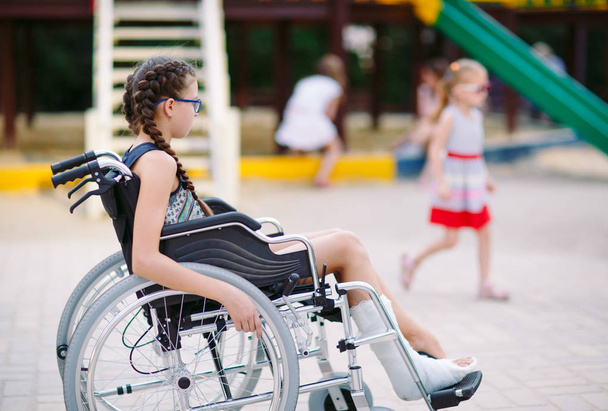 A girl with a broken leg sits in a wheelchair in front of the playground - Φωτογραφία, εικόνα