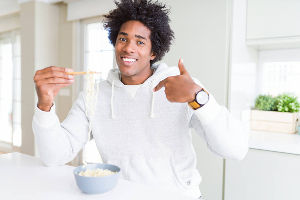 Афроамериканец ест азиатскую лапшу палочками для еды дома с удивленным лицом, указывающим пальцем на себя
 - Фото, изображение