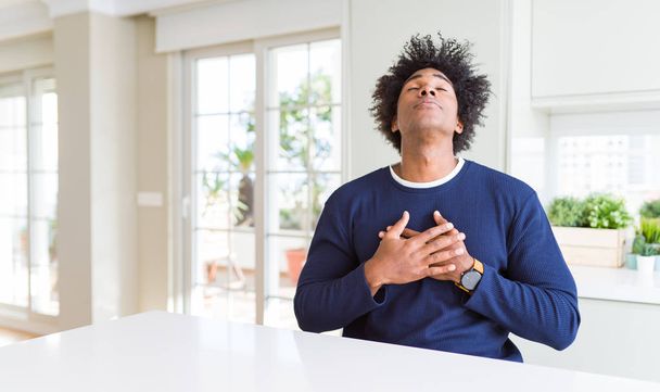 Νέος Αφρικανός Αμερικανός άνθρωπος φορώντας casual πουλόβερ κάθεται στο σπίτι χαμογελώντας με τα χέρια στο στήθος με κλειστά μάτια και ευγνωμοσύνη χειρονομία στο πρόσωπο. Έννοια της υγείας. - Φωτογραφία, εικόνα