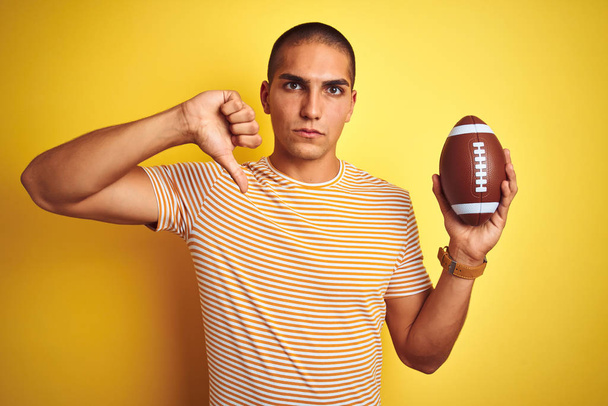 junger Rugbyspieler, der einen Fußballball über gelbem, isoliertem Hintergrund mit wütendem Gesicht hält, negatives Zeichen für Abneigung mit gesenktem Daumen, Ablehnungskonzept - Foto, Bild
