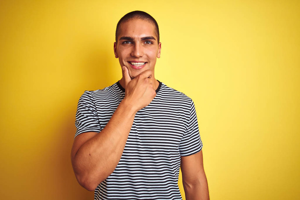 Homem bonito jovem vestindo t-shirt listrada sobre fundo isolado amarelo olhando confiante para a câmera com sorriso com braços cruzados e mão levantada no queixo. Pensar positivo
. - Foto, Imagem