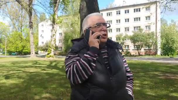 grey haired man talks on smartphone walking in city park - Video