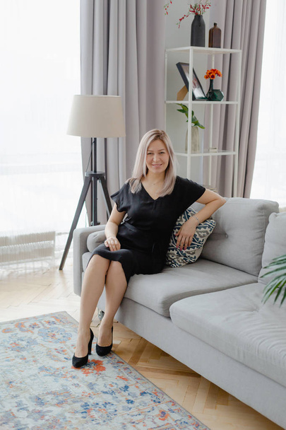 Blonde woman in a black dress sits on the couch - Fotografie, Obrázek