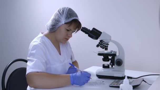Laboratorio teknikko tutkii mikroskooppi ja kirjoittaa tiedot
 - Materiaali, video