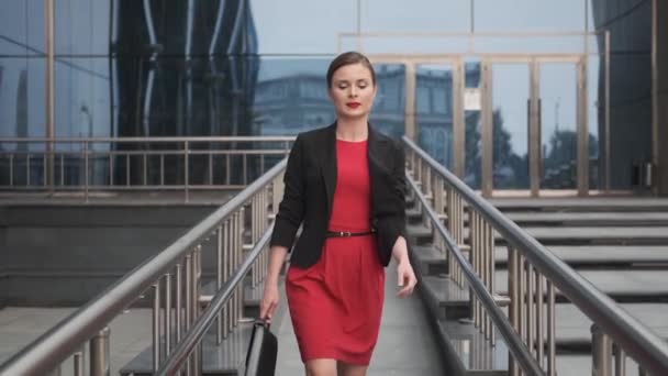 Mulher de negócios auto-confiante em um vestido vermelho andando ao lado de um centro de negócios. jovem mulher em um vestido vermelho com uma pasta em suas mãos
. - Filmagem, Vídeo