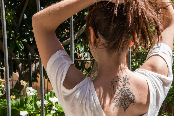 Una joven turista pelirroja con un tatuaje de Arum-lily en la espalda mira un jardín con las mismas flores en Cáceres
 - Foto, imagen