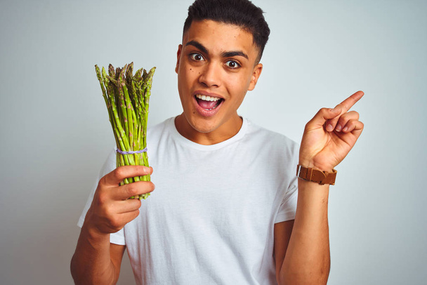 Young brazilian man eating asparagus standing over isolated grey background very happy pointing with hand and finger to the side - Φωτογραφία, εικόνα