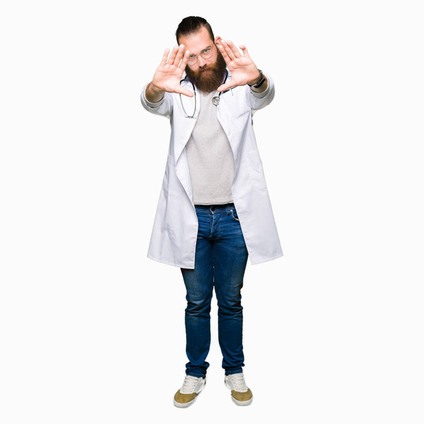 Mladá blonďatá lékař muž s vousy nosit lékařské kabát s úsměvem dělá snímek pomocí rukou dlaní a prstů, perspektivy kamery - Fotografie, Obrázek