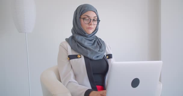 Closeup portrait of young muslim successful female doctor in hijab and eyeglasses using the laptop looking at camera sitting in the armchair in the white room indoors - Πλάνα, βίντεο