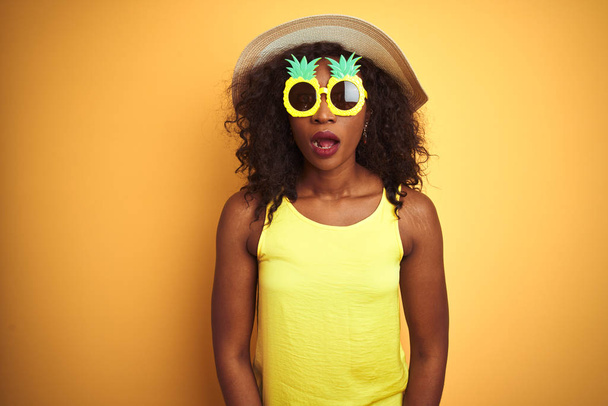 Mulher afro-americana usando óculos de sol engraçados de abacaxi sobre fundo amarelo isolado com medo e chocado com expressão surpresa, medo e rosto animado
. - Foto, Imagem