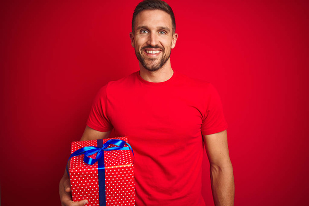 junger Mann mit Geburtstagsgeschenk auf isoliertem rotem Hintergrund mit einem glücklichen Gesicht stehend und lächelnd mit einem selbstbewussten Lächeln, das Zähne zeigt - Foto, Bild