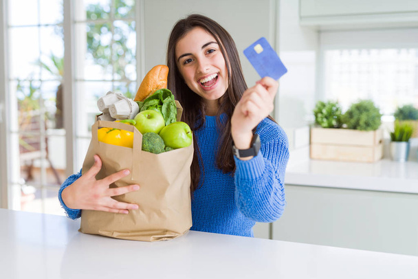 Jonge vrouw met een papieren zak vol verse boodschappen en het tonen van creditcard als betaling metod - Foto, afbeelding