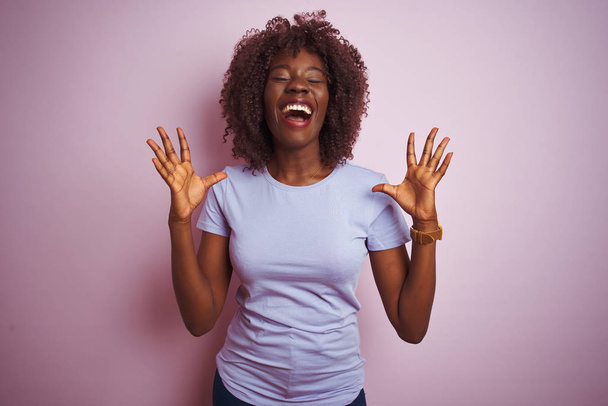 Młoda Afrykańska Afro kobieta ubrana w t-shirt stojący nad białym różowym tle obchodzi szalony i szalony na sukces z ramionami podniesione i zamknięte oczy krzyczeć podekscytowany. Koncepcja zwycięzcy - Zdjęcie, obraz