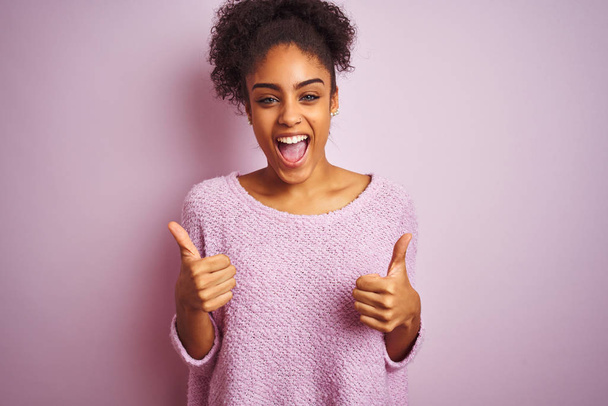 Jonge Afro-Amerikaanse vrouw dragen winter trui staande over geïsoleerde roze achtergrond succes teken doen positieve gebaar met de hand, duimen omhoog glimlachen en gelukkig. Vrolijke uitdrukking en winnaar gebaar. - Foto, afbeelding