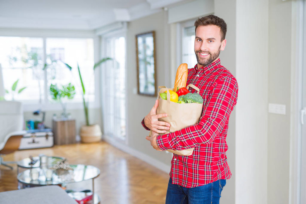 Handsome man holding paper bag full of fresh groceries at home - Фото, зображення
