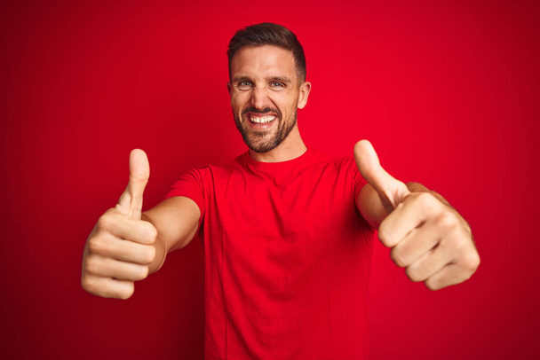 Kırmızı izole arka plan üzerinde rahat t-shirt giyen genç yakışıklı adam el ile olumlu jest yapıyor onaylanır, gülümseyen ve başarı için mutlu kadar başparmak. Kazanan jest. - Fotoğraf, Görsel