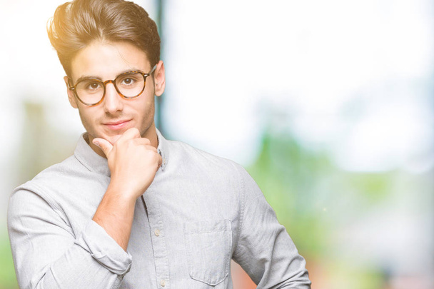 Young handsome man wearing glasses over isolated background looking confident at the camera with smile with crossed arms and hand raised on chin. Thinking positive. - Фото, изображение