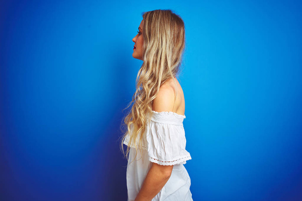 Молода красива жінка в білій сукні стоїть над синім ізольованим тлом, дивлячись в бік, розслабляє профільну позу з природним обличчям з впевненою посмішкою
. - Фото, зображення