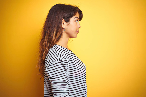 Молодая красивая женщина в полосах футболка стоя над желтым изолированным фоном глядя в сторону, расслабиться профиль позировать с естественным лицом с уверенной улыбкой
. - Фото, изображение