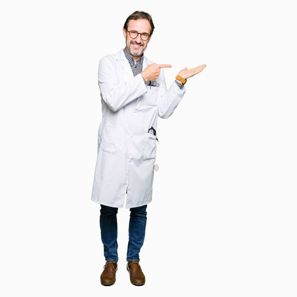 Muži středního věku lékař nosit lékařské kabát ohromen a usmívající se na kameru při prezentaci s ruku a ukazoval prstem. - Fotografie, Obrázek
