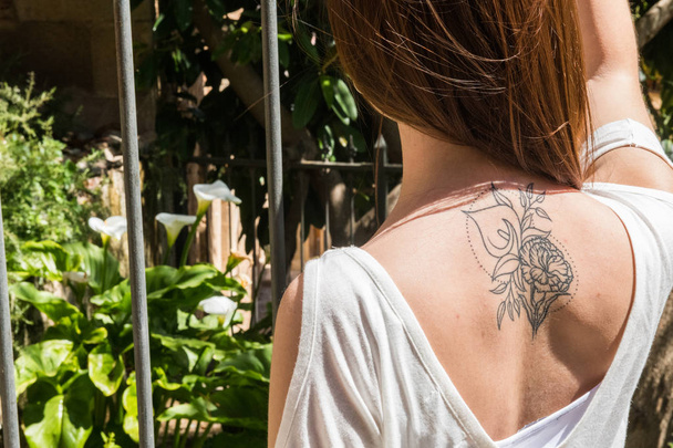 Uma jovem turista ruiva com uma tatuagem de lírio-arum nas costas olha para um jardim com as mesmas flores em Cáceres.
 - Foto, Imagem