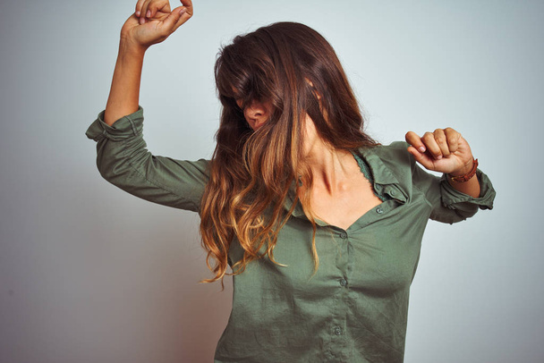 Молодая красивая женщина в зеленой рубашке, стоящей на сером изолированном фоне, танцует счастливой и веселой, улыбаясь, двигаясь небрежно и уверенно слушая музыку
 - Фото, изображение
