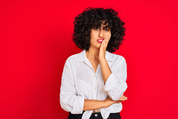 Young arab woman with curly hair wearing white casual shirt over isolated red background thinking looking tired and bored with depression problems with crossed arms. - Foto, afbeelding