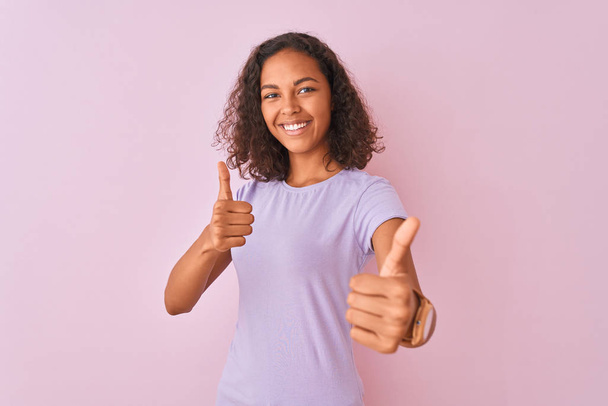 Młoda Brazylijska kobieta nosi t-shirt stojący na białym tle różowy zatwierdzania robi pozytywny gest z ręki, kciuki się uśmiechnięty i szczęśliwy dla sukcesu. Gest zwycięzcy. - Zdjęcie, obraz