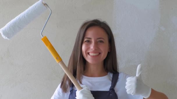 若い女の子が修理をする。ローラーを持った少女は壁にペンキを塗る。女の子は修理をして笑顔を作る。家の壁を塗る建設と修理。修理作業 - 映像、動画