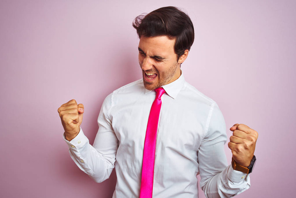 Молодой красивый бизнесмен в рубашке и галстуке стоя на изолированном розовом фоне очень счастлив и взволнован делая жест победителя с поднятыми руками, улыбаясь и крича об успехе. Концепция празднования
. - Фото, изображение