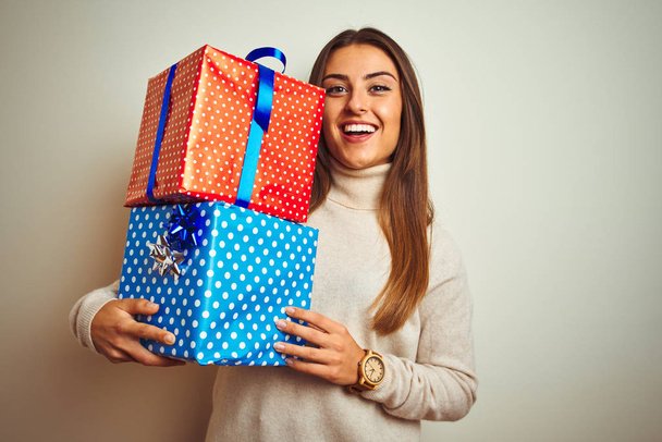 Молодая красивая женщина держит подарки на день рождения стоя на изолированном белом фоне с счастливым лицом стоя и улыбаясь с уверенной улыбкой показывая зубы
 - Фото, изображение