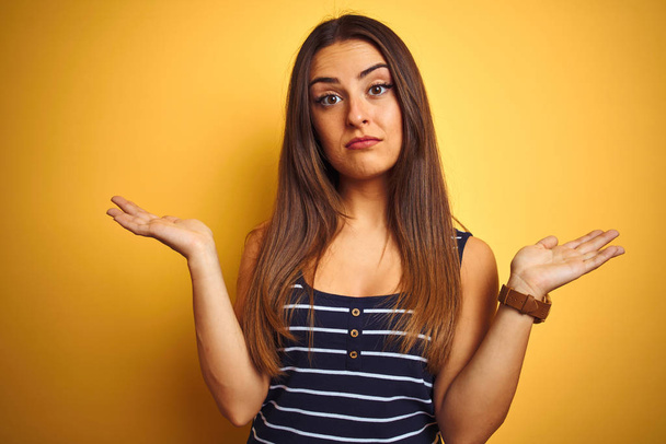 Young beautiful woman wearing striped t-shirt standing over isolated yellow background clueless and confused expression with arms and hands raised. Doubt concept. - Photo, Image