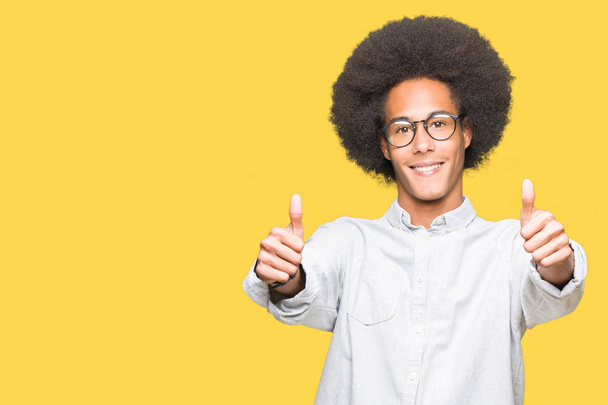 Młody african american włosów afro zatwierdzające noszenie okularów robi pozytywny gest ręką, kciuki, uśmiechając się i szczęśliwy dla sukcesu. Patrząc na kamery, zwycięzca gest. - Zdjęcie, obraz