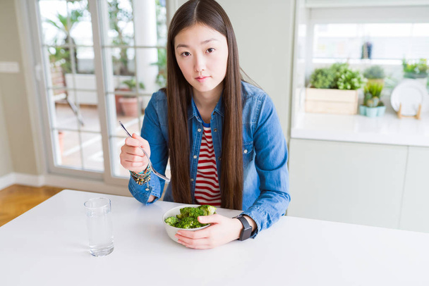 Beautiful Asian woman eating green fresh broccoli with a confident expression on smart face thinking serious - Photo, Image
