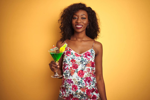 Genç afro-amerikan kadın mutlu bir yüz ayakta ve dişleri gösteren kendinden emin bir gülümseme ile gülümseyerek izole sarı arka plan üzerinde duran kokteyl içme - Fotoğraf, Görsel