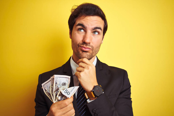 Молодий красивий бізнесмен в костюмі тримає долари над ізольованим жовтим тлом серйозне обличчя, думаючи про питання, дуже заплутана ідея
 - Фото, зображення