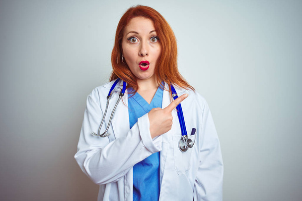 Молодая рыжая женщина врач, использующая стетоскоп на белом изолированном фоне Удивлен указывая пальцем в сторону, открытый рот поражен выражение
. - Фото, изображение