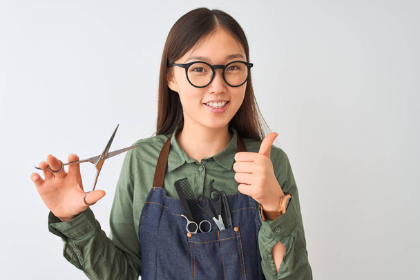 孤立した白い背景の上にハサミを持ったメガネをかけている中国の美容師女性は、OKサインをやって大きな笑顔で幸せ、指で親指を上げ、優れた兆候 - 写真・画像