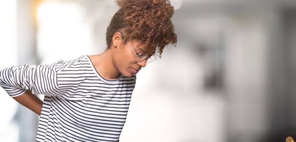 Красивая молодая африканская американка в очках на изолированном фоне, страдает от боли в спине, трогает спину рукой, мускулистая боль
 - Фото, изображение