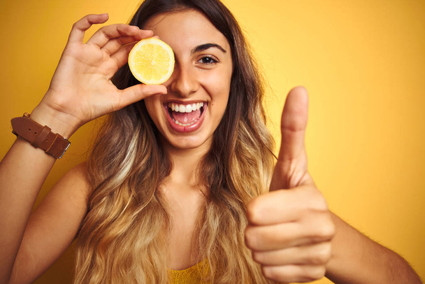 黄色の孤立した背景の上に目にレモンを保持している若い美しい女性は、OKサインをやって大きな笑顔で幸せ、指で親指アップ、優れた兆候 - 写真・画像