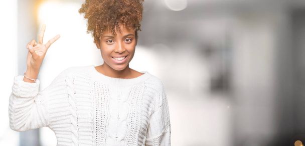 Hermosa joven afroamericana americana vistiendo suéter de invierno sobre fondo aislado mostrando y señalando con los dedos número dos mientras sonríe confiada y feliz
. - Foto, imagen
