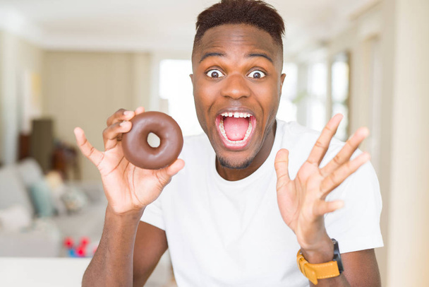 Uomo afroamericano mangiare ciambella al cioccolato molto felice ed eccitato, espressione vincitore celebrando la vittoria urlando con grande sorriso e mani alzate
 - Foto, immagini